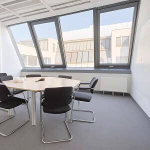 Heller Besprechungsraum mit schrägen Fenstern, einem sechseckigen Tisch und grauen Stühlen.