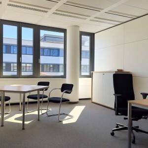 Helles Büro mit großen Fenstern, Säulen und Teppichboden, ausgestattet mit Schreibtischen.