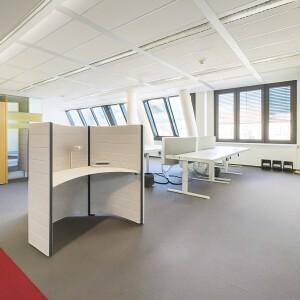 Offenes Büro mit schrägen Fenstern, modularen Arbeitsplätzen und grauem Bodenbelag.