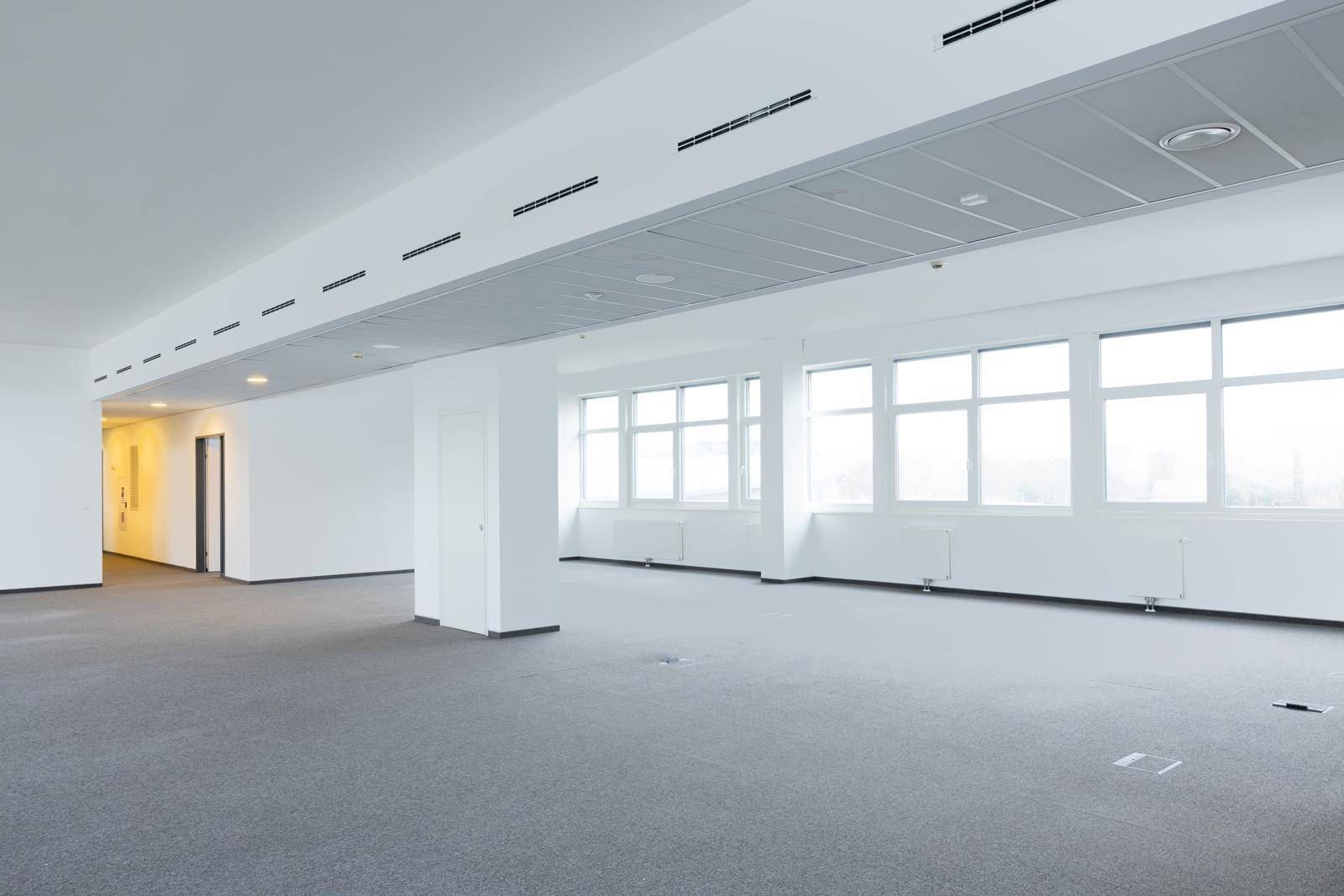 Großes, leeres Büro mit Teppichboden, vielen Fenstern und integrierter Deckenbeleuchtung und Lüftung.