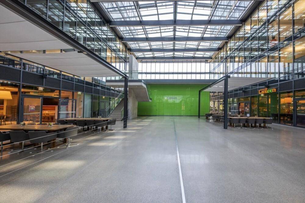 Weitläufiges Atrium mit Glasdach, Betonboden, Sitzbereichen und einer auffälligen grünen Wand im Hintergrund.