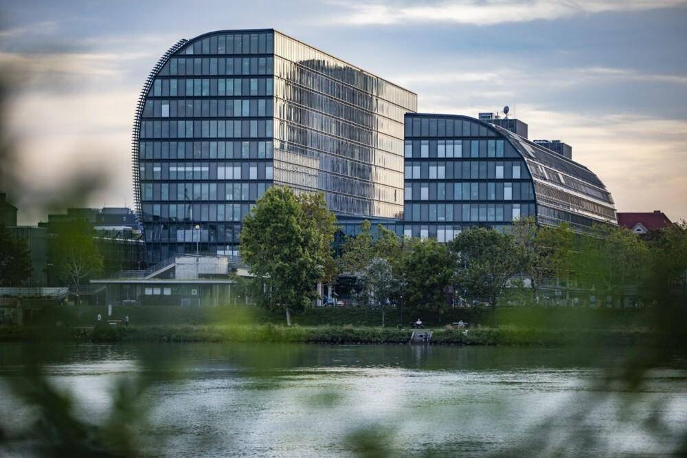 Modernes Bürogebäude mit geschwungener Glasfassade am Flussufer unter einem bewölkten Himmel.