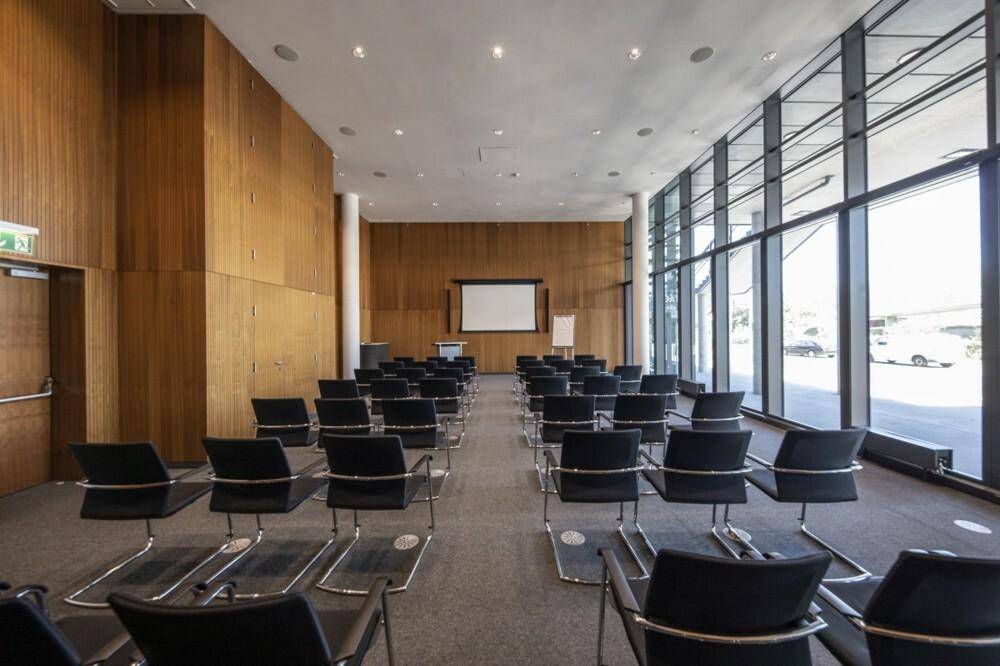 Großer Konferenzraum mit Reihen von schwarzen Stühlen, einer Projektionsfläche und Holzwänden.