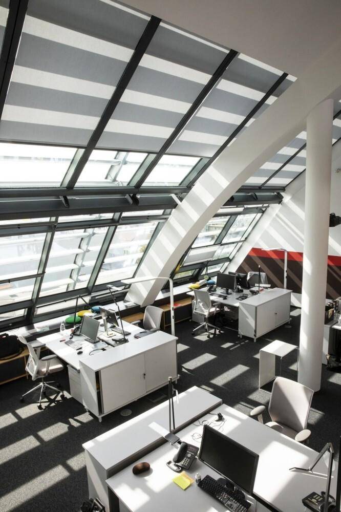 Offener Bürobereich mit schrägen Glasfenstern, mehreren Schreibtischen und Bürostühlen.