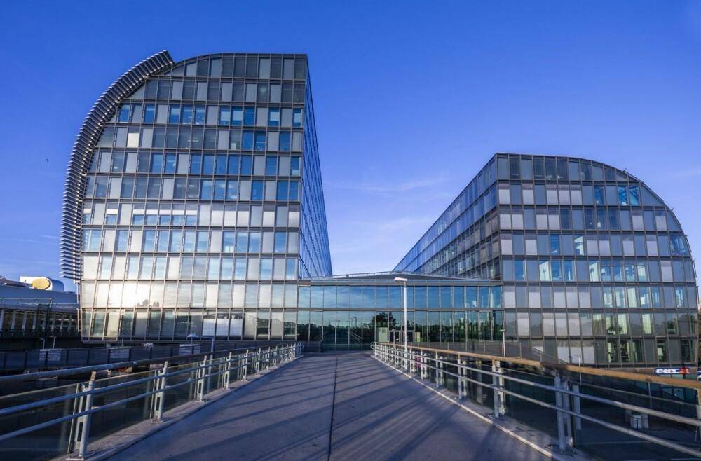 Modernes Bürogebäude mit geschwungener Glasfassade, gesehen von einer Fußgängerbrücke unter blauem Himmel.