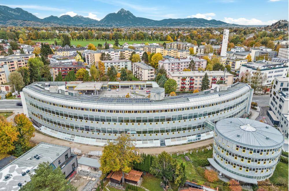 Luftaufnahme eines großen, geschwungenen Bürogebäudes in einer städtischen Umgebung mit Bergen im Hintergrund.