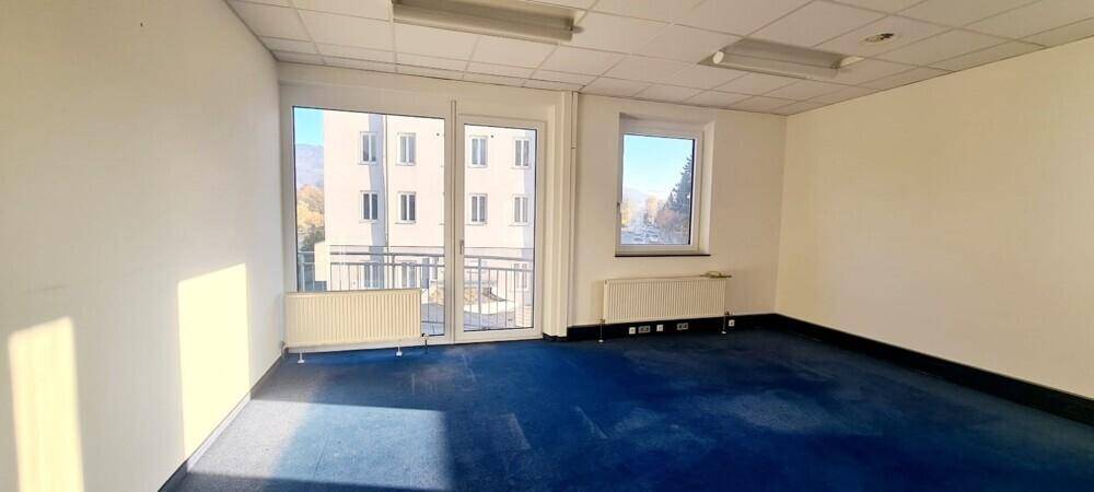 Leeres Büro mit blauem Teppichboden, großen Fenstern und Zugang zu einem Außenbereich.