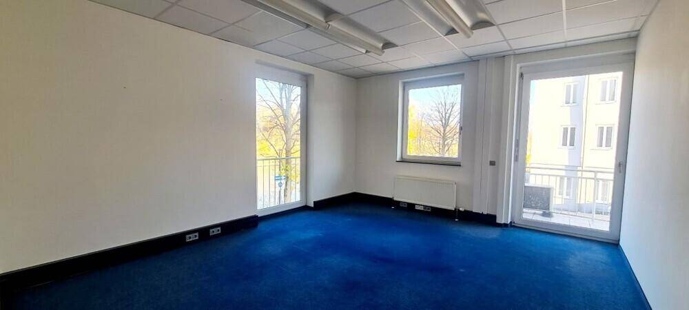 Helles Büro mit blauem Teppichboden, Heizkörpern und Zugang zu einem kleinen Balkon.