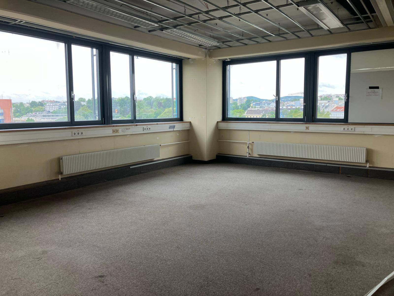 Leeres Büro mit Teppichboden, großen Fenstern und Heizkörpern, bietet einen weiten Blick auf die Stadt.