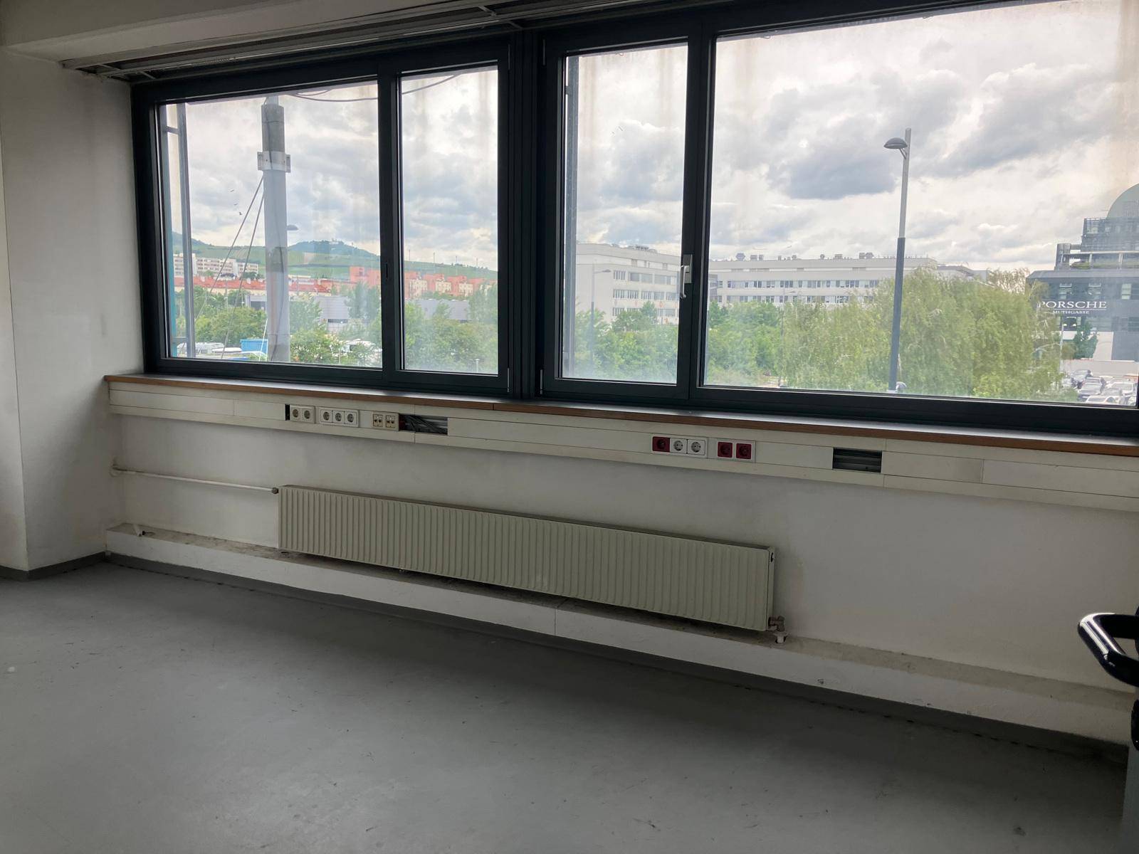 Leerer Raum mit großen Fenstern, die einen Blick auf die Stadt bieten, und einem langen Heizkörper darunter.