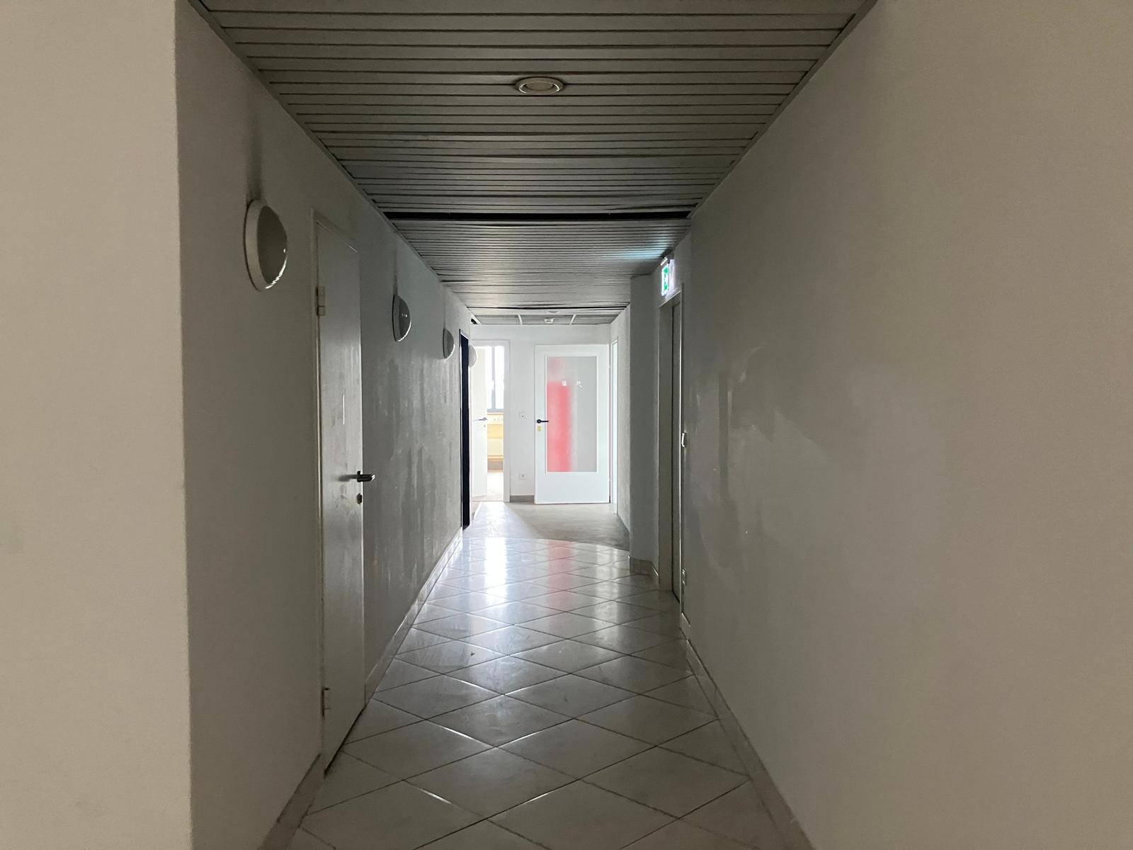 Heller Flur mit weißen Wänden und diagonal verlegten Fliesen, der zu mehreren Türen führt.