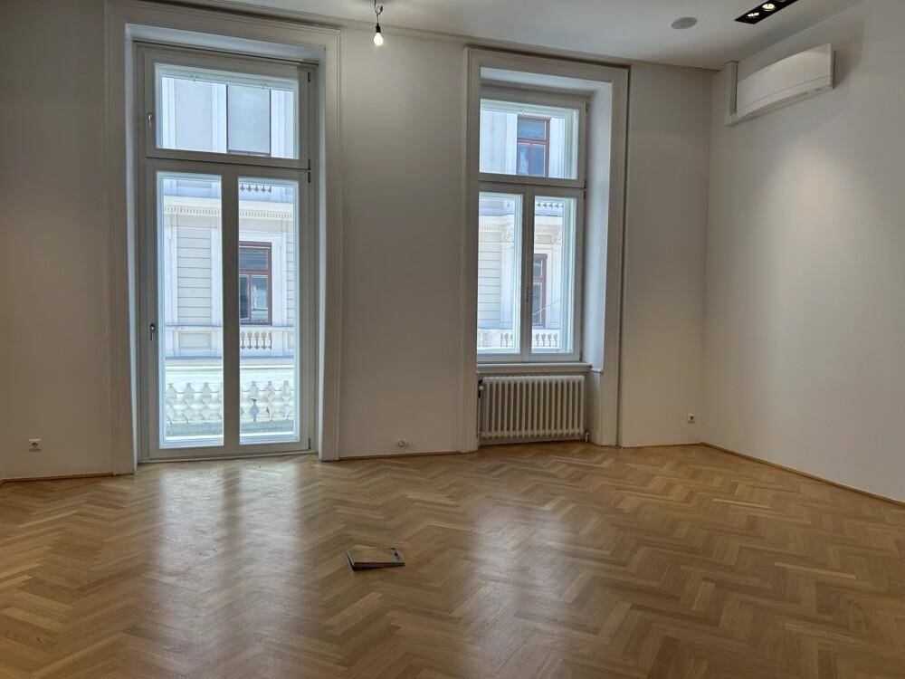 Geräumiger Raum mit Fischgrätparkett, hohen Fenstern und Klimaanlage, bietet Zugang zu einem französischen Balkon.