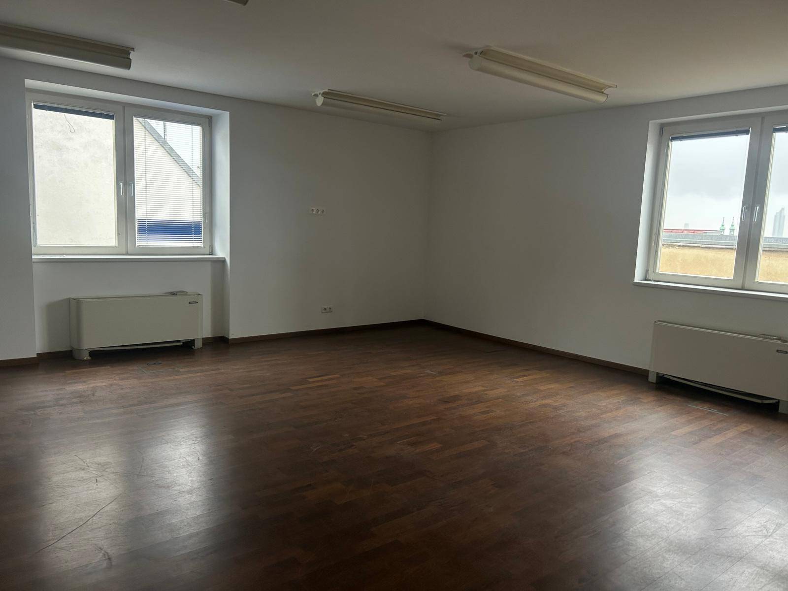 Geräumiger Büroraum mit dunklem Holzboden und großen Fenstern, die viel Tageslicht hereinlassen.