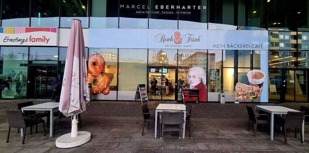 Außenbereich eines Cafés mit Tischen, Stühlen und Sonnenschirm vor einer modernen Geschäftsfassade.