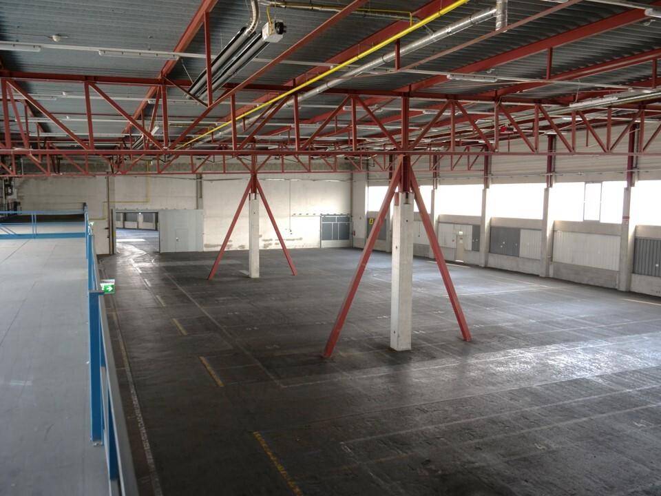 Große Lagerhalle mit Betonboden, roten Stahlstützen und einem blauen Geländer, das eine erhöhte Ebene andeutet.