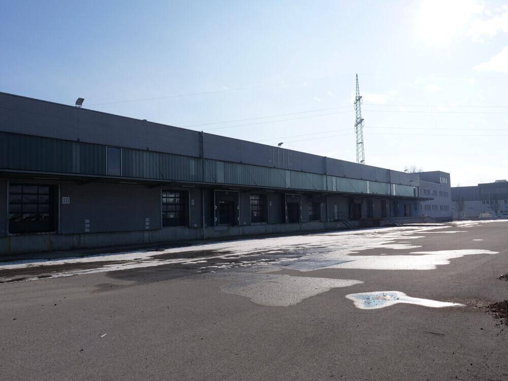 Lange Ansicht des Industriegebäudes mit zahlreichen Laderampen und einem weitläufigen asphaltierten Vorplatz unter blauem Himmel.