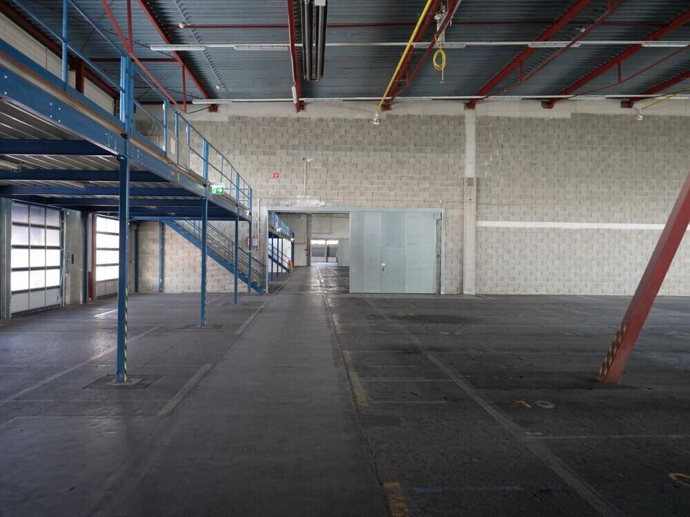 Geräumige Lagerhalle mit hoher Decke, Betonboden und einer blauen Stahlkonstruktion mit Treppenaufgang.