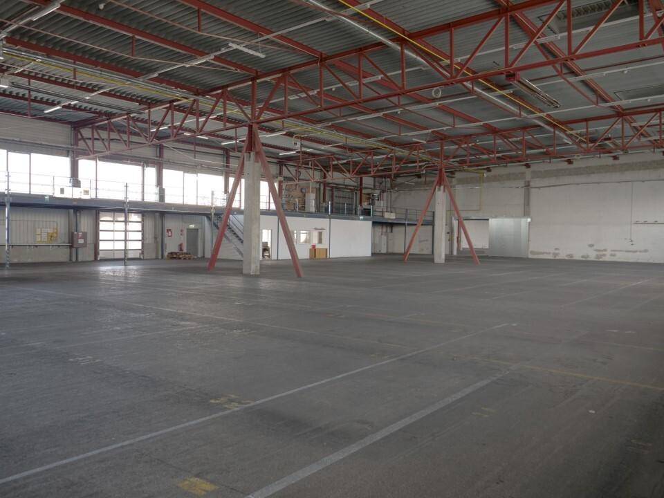 Weitläufige Lagerhalle mit hohen Decken, Betonboden und roten Stahlkonstruktionen, inklusive kleiner Büroeinheit.
