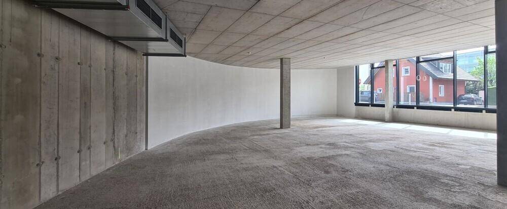 Unfertiger Gewerberaum mit geschwungener Wand, Betonoberflächen und großen Fenstern mit Blick auf ein Gebäude.