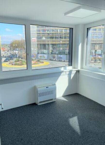 Ecke eines hellen Büroraums mit großen Fenstern, die einen Blick auf die Stadt und eine Kreuzung bieten.