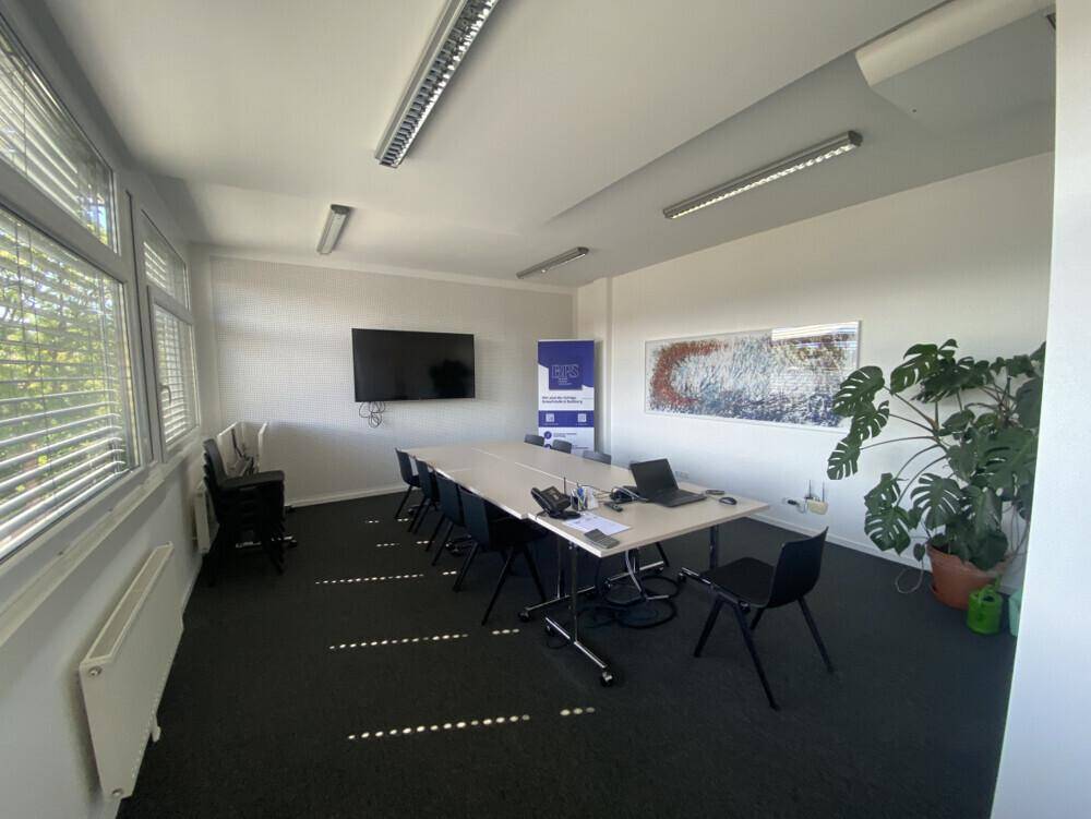 Geräumiger Konferenzraum mit langem Tisch, vielen Stühlen, Wandfernseher und großen Fenstern.