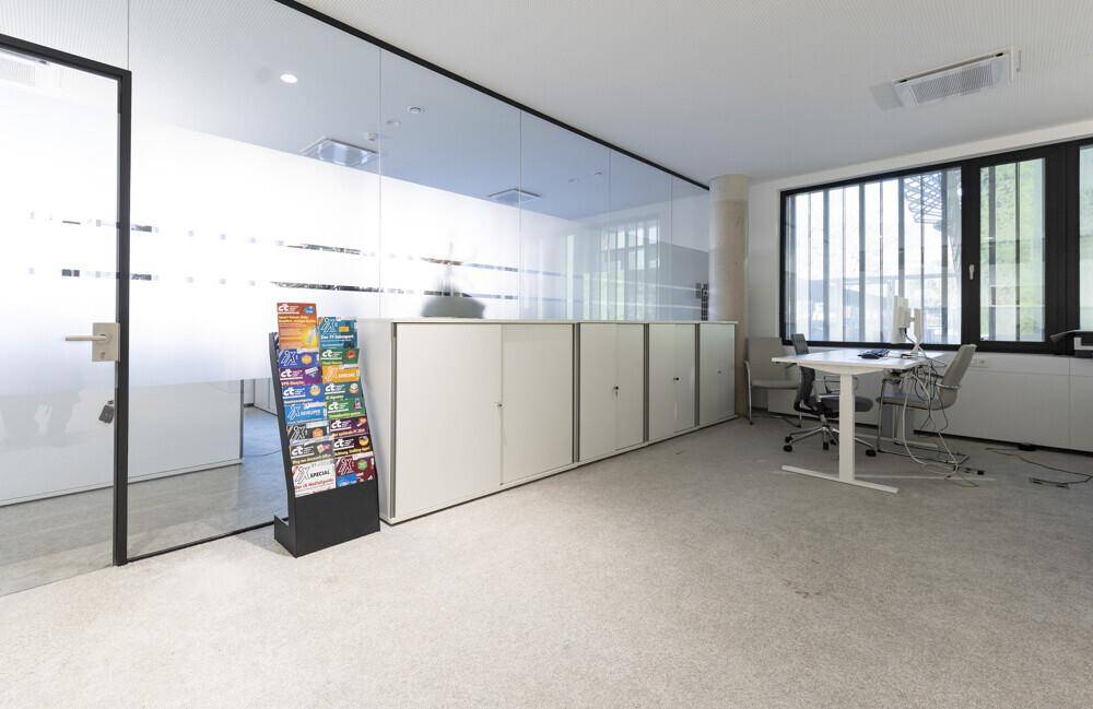 Moderner Bürobereich mit Glaswänden, Teppichboden und einem Zeitschriftenständer.