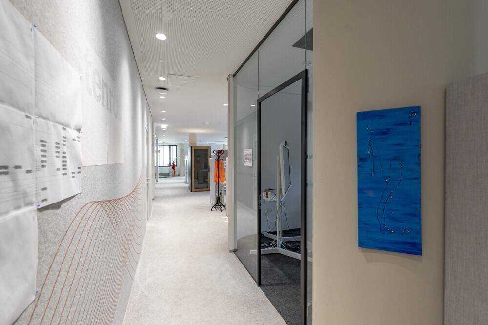 Heller Bürogang mit Teppichboden, Glaswänden und moderner Kunst an der Wand.