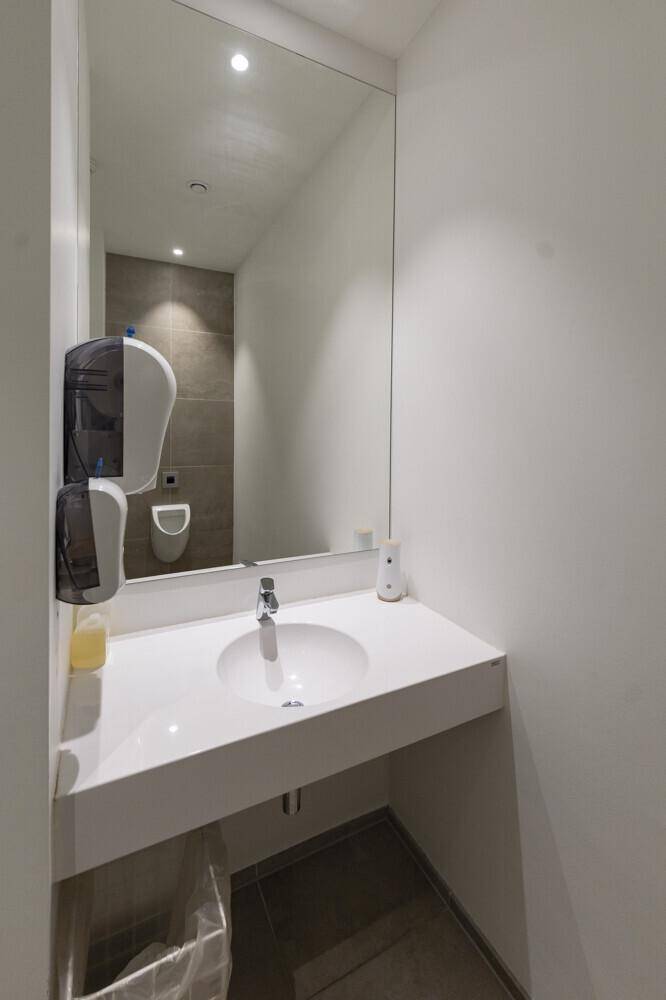 Modernes Badezimmer mit weißem Waschtisch, großem Spiegel und Urinal im Hintergrund.