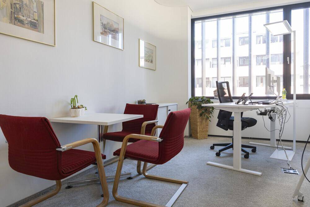 Helles Einzelbüro mit Schreibtisch, Besprechungstisch und großen Fenstern.