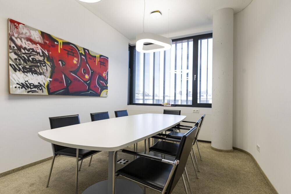 Besprechungsraum mit einem ovalen weißen Tisch, schwarzen Stühlen und einem farbenfrohen Kunstwerk an der Wand.