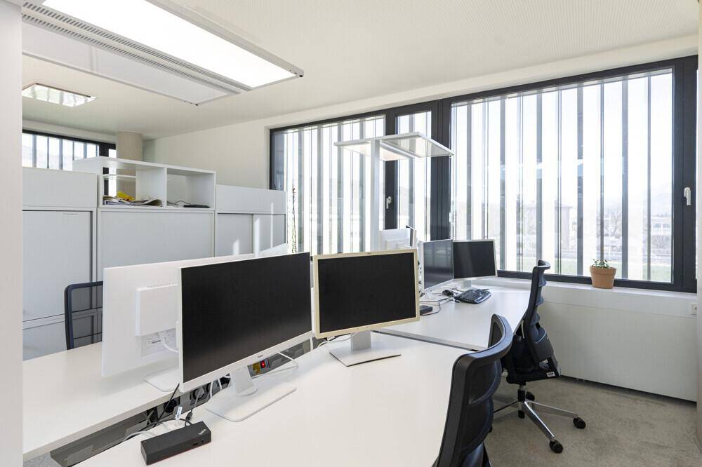 Moderner Arbeitsplatz mit zwei Monitoren, viel Tageslicht und Blick auf die Umgebung durch große Fenster.