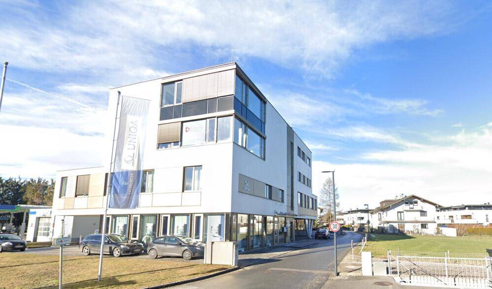 Modernes Geschäftsgebäude mit mehreren Etagen, großen Fenstern und geparkten Autos davor.