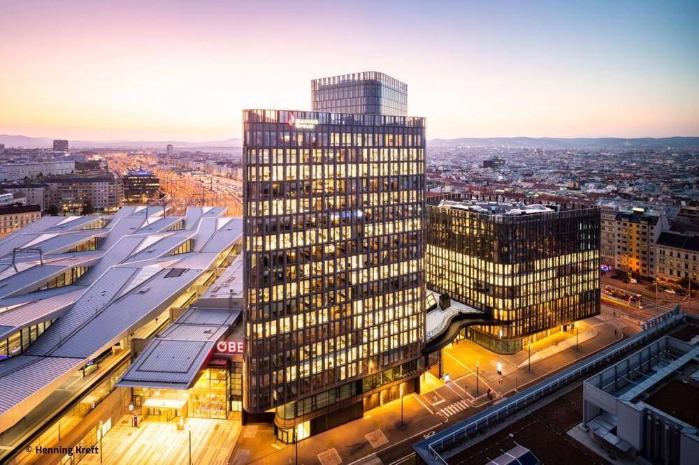 Luftaufnahme moderner Gebäude und eines Bahnhofs mit Stadtpanorama bei Sonnenuntergang.