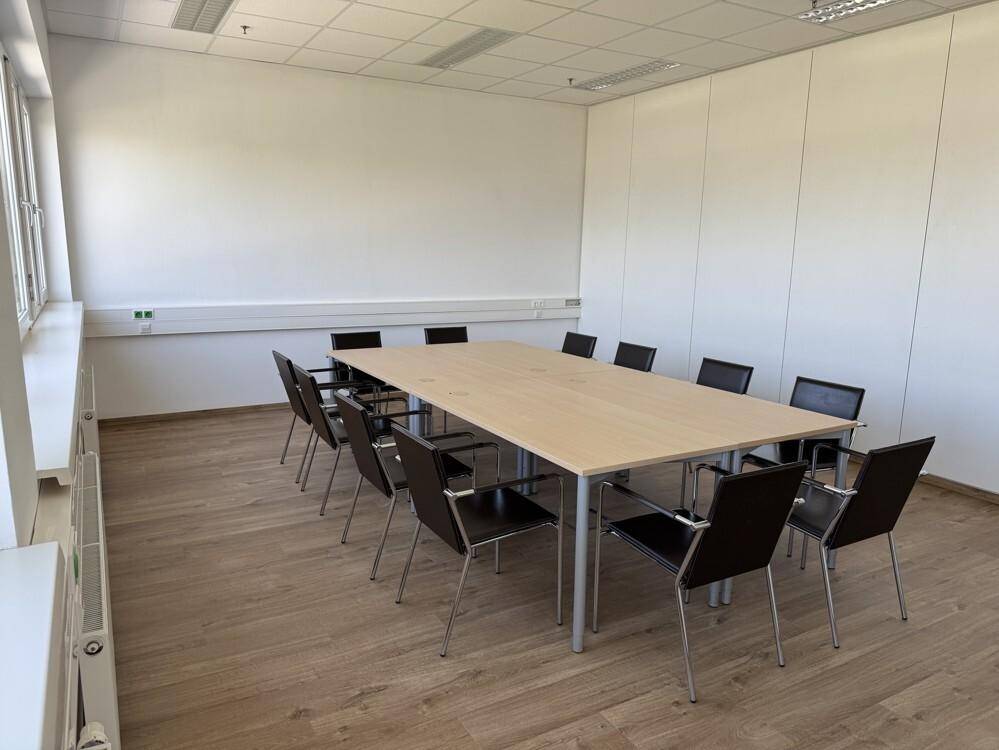 Heller Konferenzraum mit großem Holztisch, schwarzen Stühlen und Laminatboden.