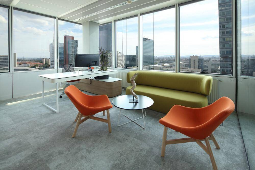 Modernes Büro mit Schreibtisch, grüner Couch und orangefarbenen Stühlen vor großen Fenstern mit Stadtblick.