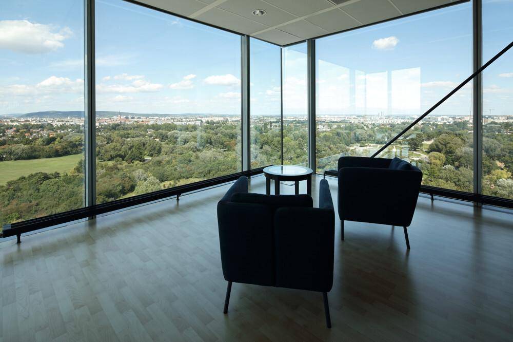 Heller Büroraum mit zwei blauen Sesseln, kleinem Tisch und Panoramafenstern mit Stadt- und Grünblick.