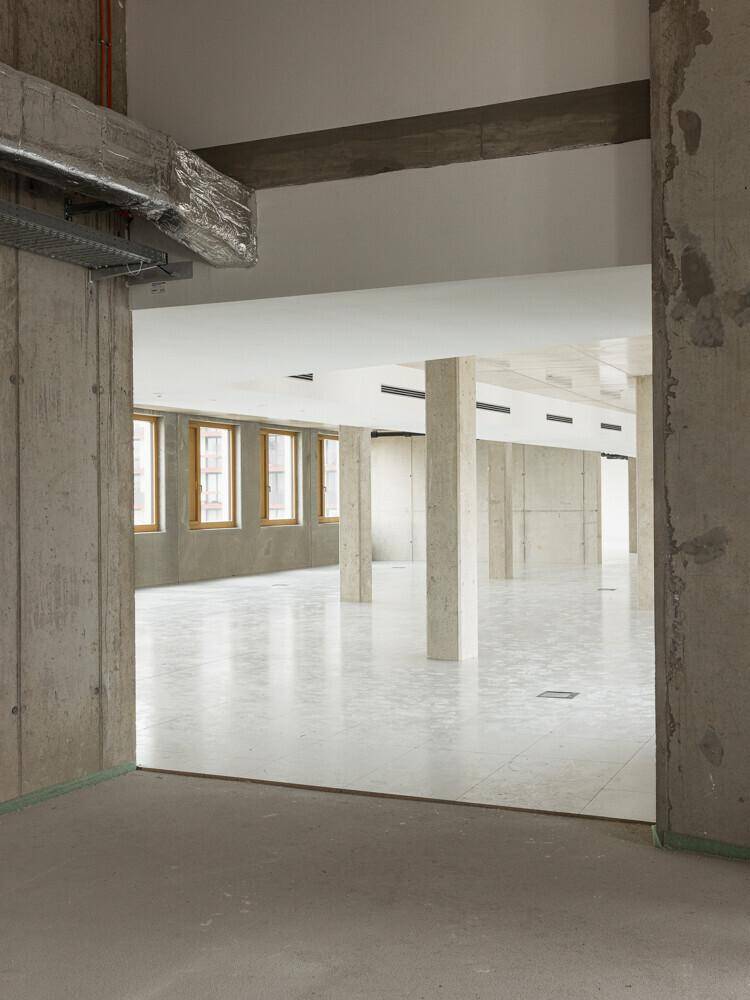 Blick von einem Rohbaubereich in einen größeren, offenen Raum mit Betonstützen und Fliesenboden.