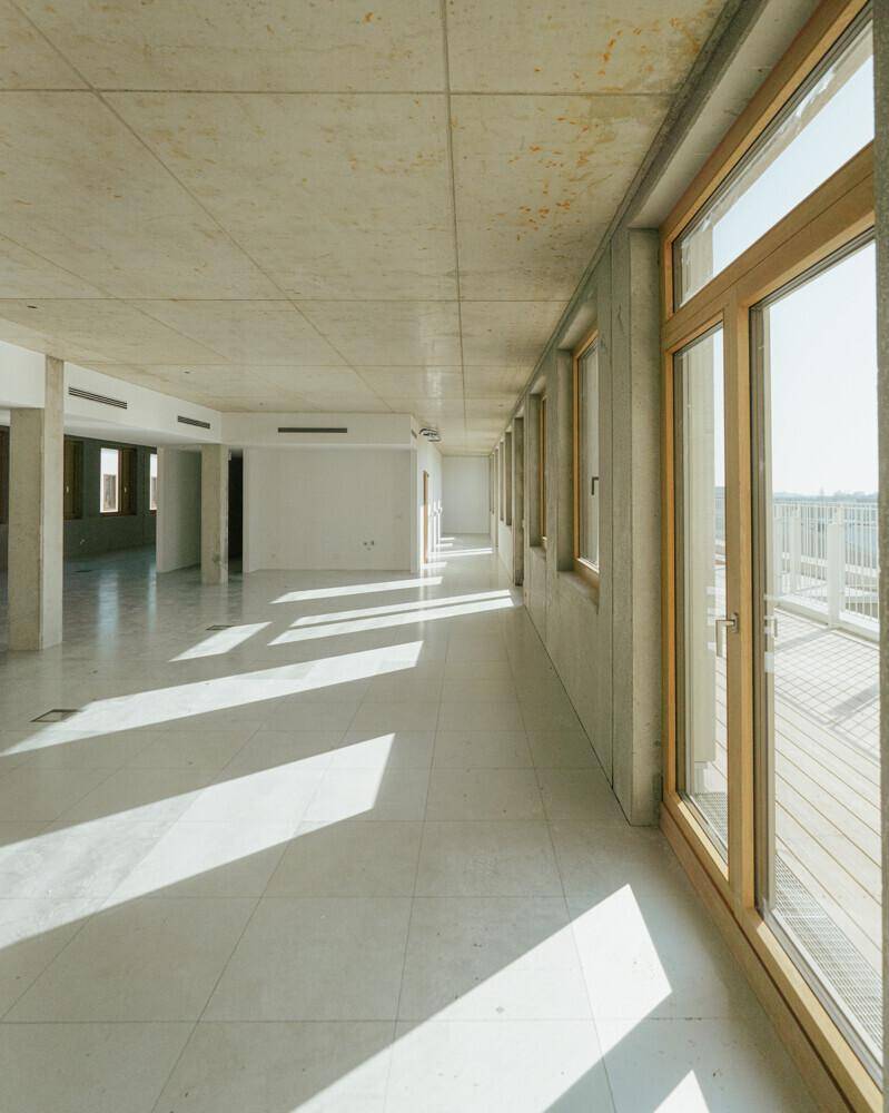 Langer, heller Raum im Rohbau mit Fliesenboden und einer Reihe von Fenstern, die Tageslicht hereinlassen.
