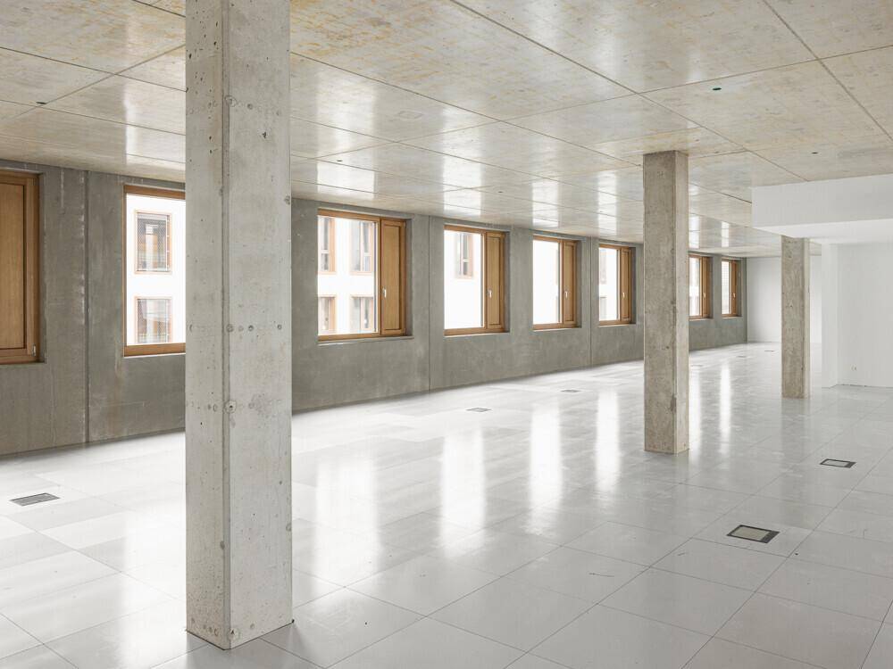 Weitläufiger Rohbau mit Betonstützen, glänzendem Fliesenboden und Holzfenstern, die viel Licht spenden.