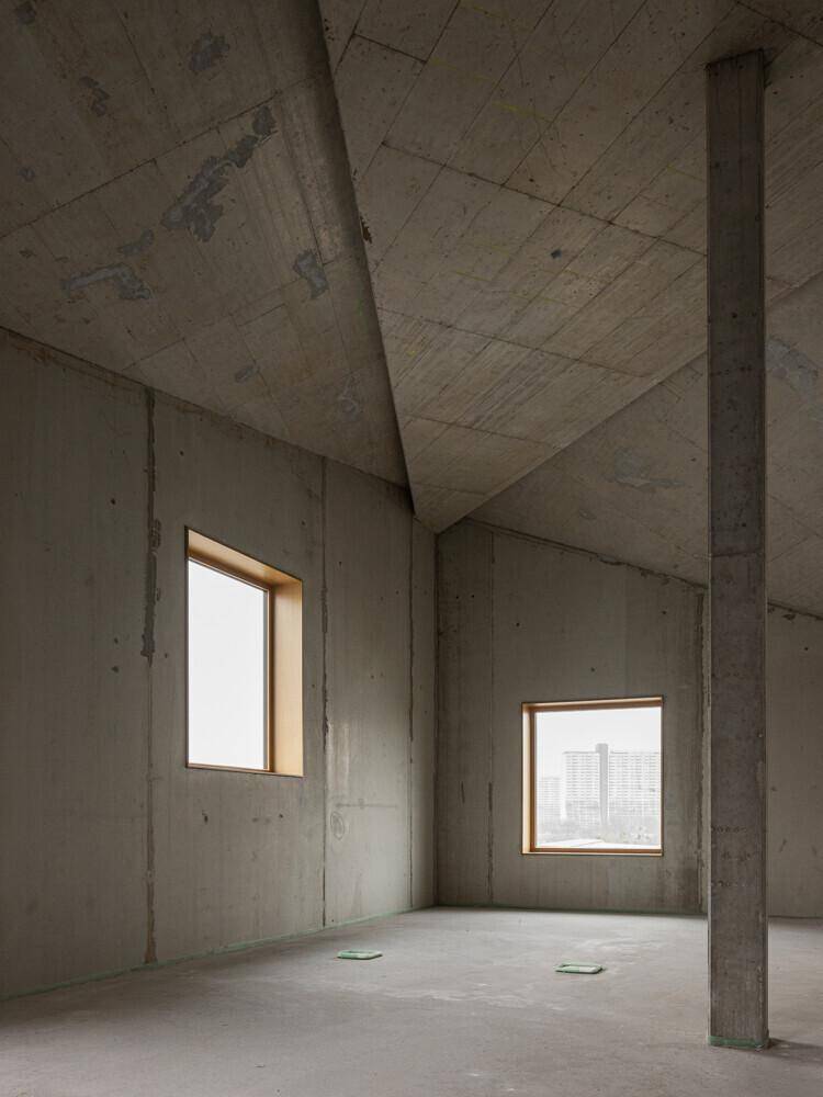 Rohbau mit schrägen Betonwänden und -decken sowie kleinen Fenstern, die Tageslicht hereinlassen.