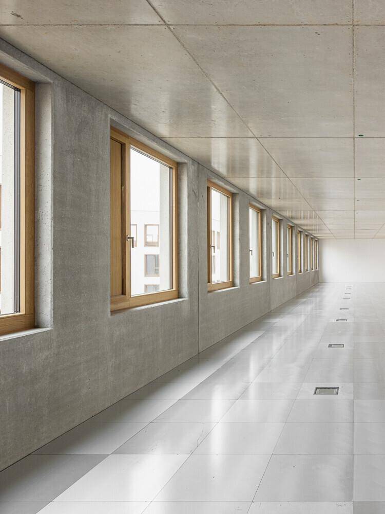 Langer, leerer Raum im Rohbau mit Betonwänden, Fliesenboden und einer Reihe von Holzfenstern.