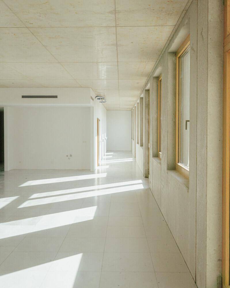 Unfertiger Innenraum mit weißem Fliesenboden, Betonwänden und Fenstern, die viel Sonnenlicht hereinlassen.