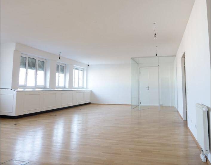 Heller, leerer Raum mit Holzboden, großen Fenstern und einer modernen Glaswand als Raumteiler.