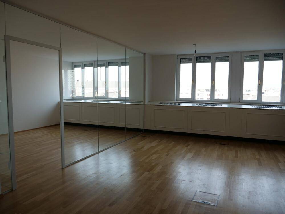 Großer, heller Raum mit Holzboden, einer verspiegelten Wand und Fenstern mit Rollläden.