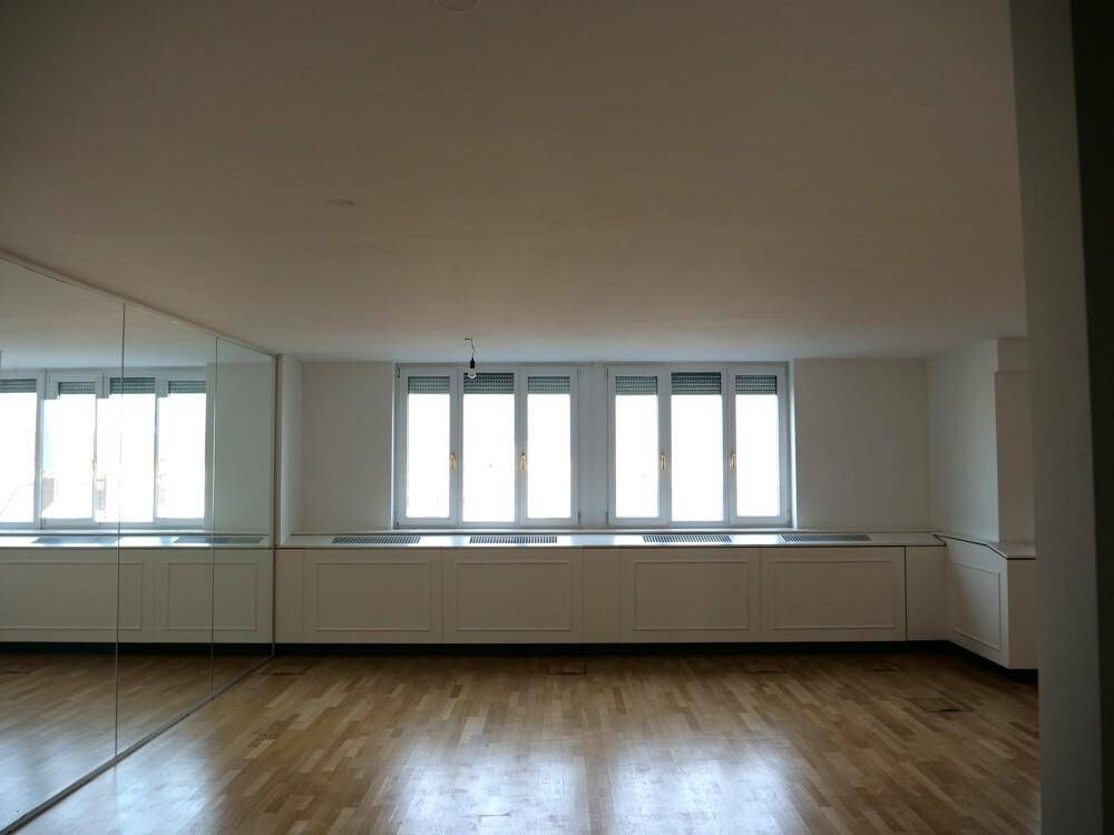 Heller, leerer Raum mit Holzboden und einer Reihe großer Fenster, die viel Tageslicht hereinlassen.