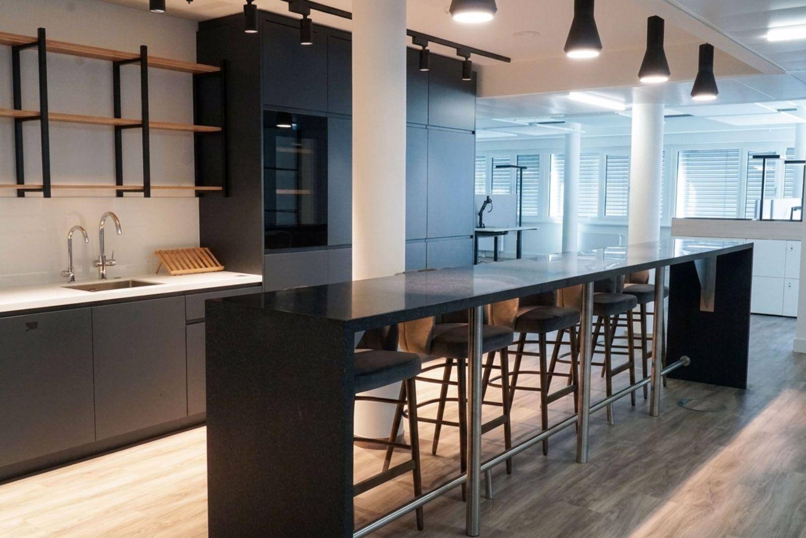 Elegante Büroküche mit dunklen Schränken, langer Bar und offenen Holzregalen unter moderner Beleuchtung.
