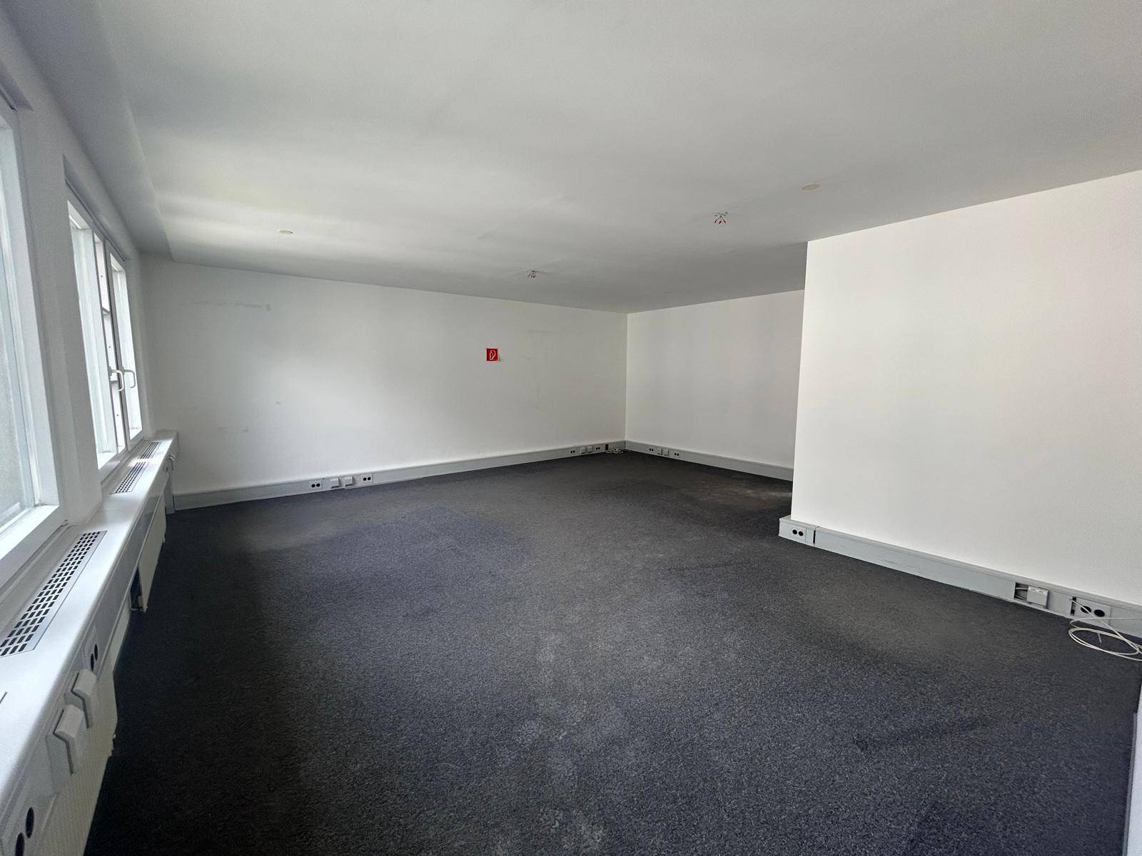 Geräumiges, leeres Büro mit Teppichboden und großen Fenstern, die für eine helle Arbeitsatmosphäre sorgen.