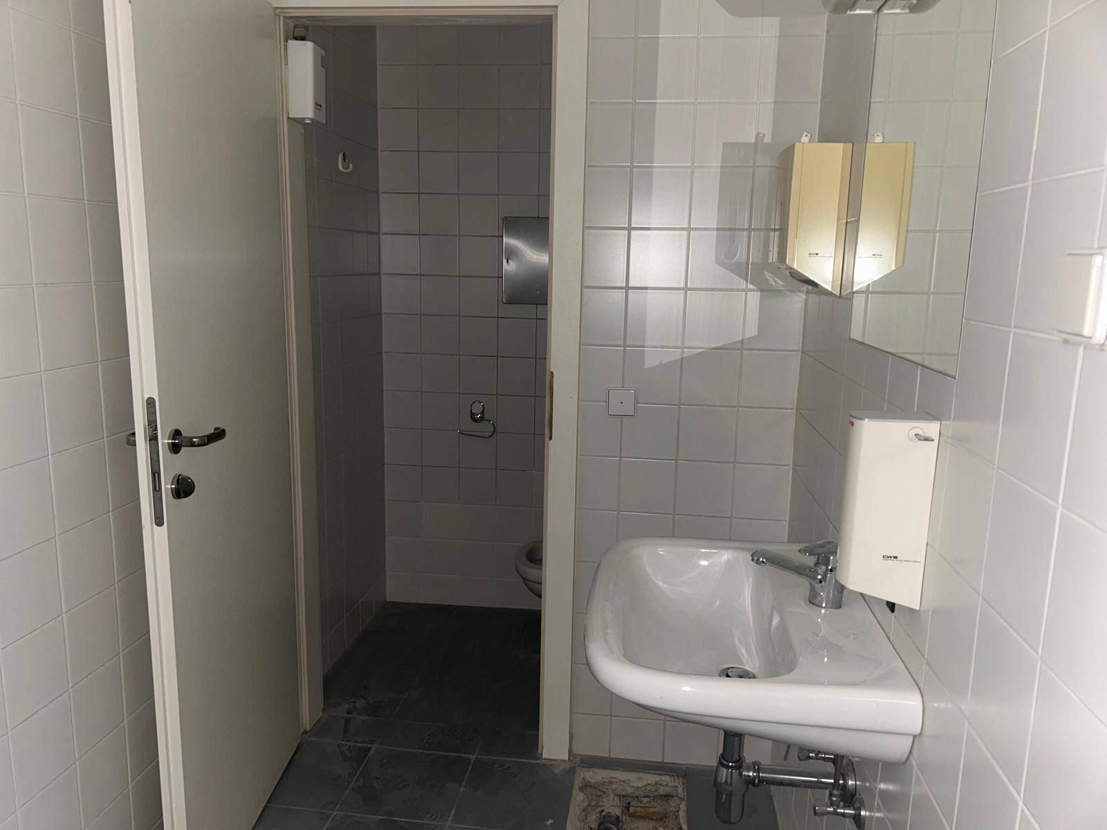 Einfacher Toilettenraum mit weißen Fliesen, einem Waschbecken und einer Toilette im Hintergrund.