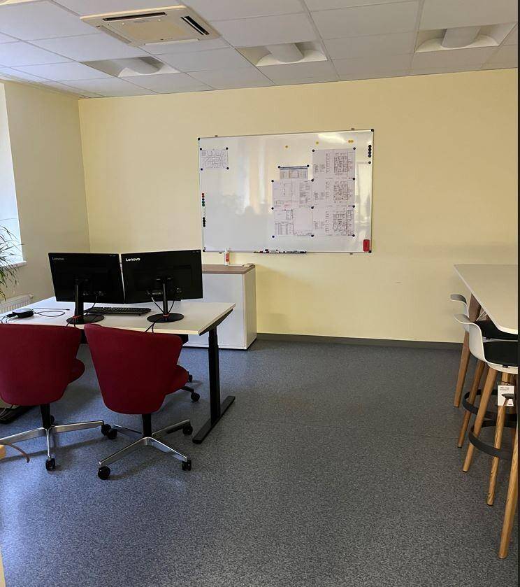 Heller Büroraum mit zwei Arbeitsplätzen, roten Stühlen und einem Whiteboard an der gelben Wand.