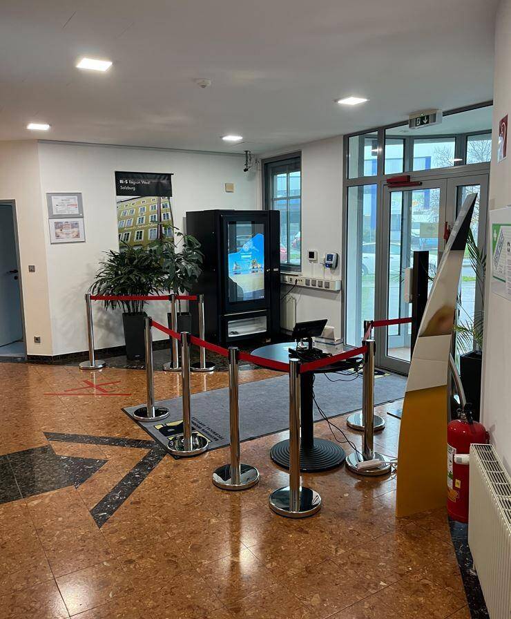 Empfangsbereich mit dunklem Terrazzoboden, Absperrungen und einem Informationsbildschirm.